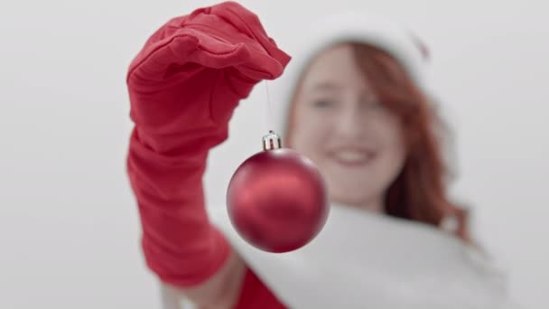 Прикраса ялинки, червона куля в руках дівчини, що тримає її в червоних рукавичках, вказує на камеру перед нею і посміхається. Концепція різдвяних та новорічних свят. Висока якість - Кадри, відео