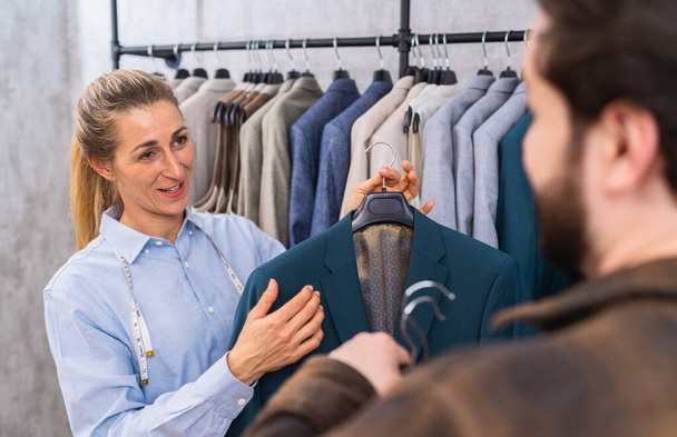 Schneider hält für einen männlichen Kunden eine Jacke in Kricketblau, beide unterhalten sich in einem Geschäft - Foto, Bild