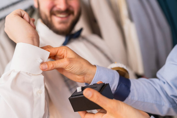 Nahaufnahme eines maßgeschneiderten Manschettenknöpfchens auf dem Hemdsärmel eines Kunden, beide lächeln in einem Hochzeitsgeschäft - Foto, Bild
