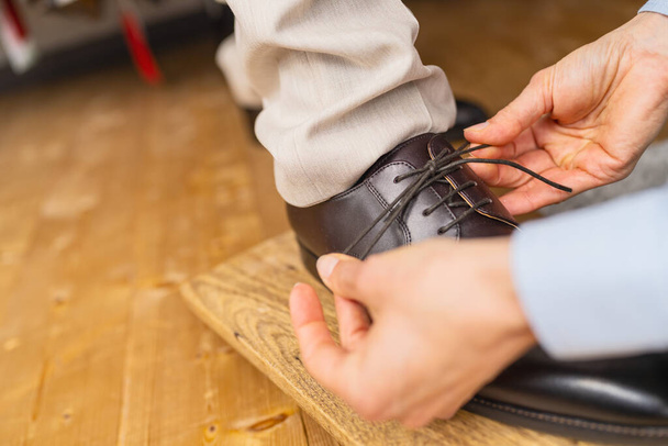 Крупный план рук, завязывающих шнурки новой коричневой кожаной туфли на деревянной доске в свадебном магазине - Фото, изображение