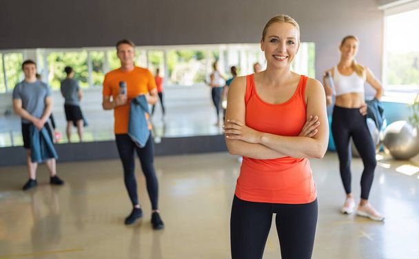 Mujer sonriente en camiseta naranja con los brazos cruzados en un gimnasio, gente en el fondo. Trabajo en equipo Concepto imagen - Foto, imagen