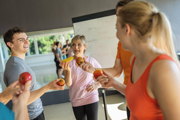 Ομάδα γυμναστικής συζητά τη διατροφή κρατώντας φρούτα με ένα λευκό πίνακα στο παρασκήνιο - Φωτογραφία, εικόνα