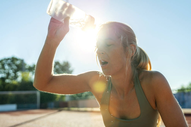 Kobieta wylewa wodę na twarz po treningu, rozbłysk słońca, buteleczka sportowa, orzeźwiająca - Zdjęcie, obraz