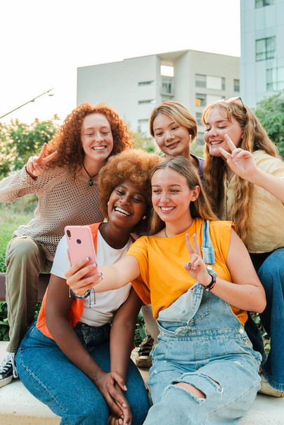 Pionowe. Szczęśliwe wielorasowe młode kobiety patrzące w kamerę ze smartfonem cieszące się razem siedząc na zewnątrz. Zabawne panie bawią się kręcąc selfie portret swoich przyjaciół z telefonu komórkowego. Wysoka - Zdjęcie, obraz