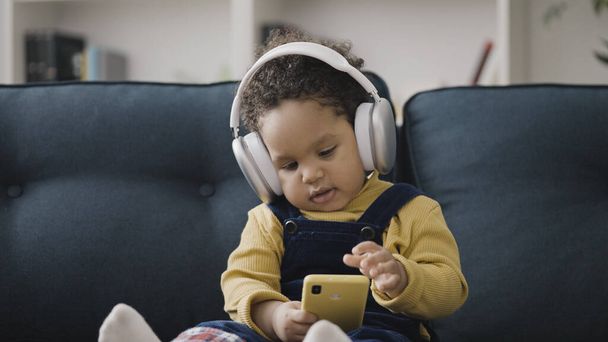 Cute toddler wearing large headphones, sitting on sofa and playing on smartphone - Zdjęcie, obraz