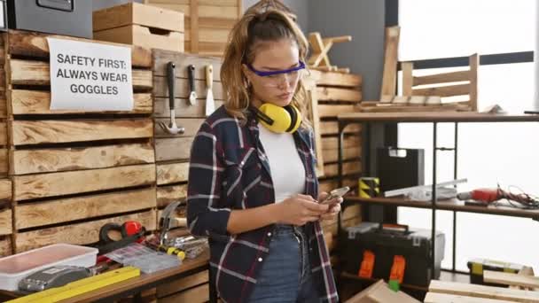 Sonriente joven mujer hispana carpintera alegremente enviando mensajes de texto a su negocio de carpintería, balanceando con confianza sus gafas de seguridad - Imágenes, Vídeo