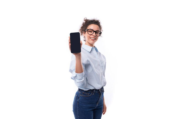 портрет молодой умной брюнетки-ассистентки, одетой в простую синюю рубашку и джинсы, держащей смартфон с экраном впереди нее. - Фото, изображение