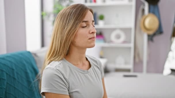 Aantrekkelijke jonge blonde vrouw, mediteren terwijl rustig zitten op de thuisbank, lucht inademen om angst in evenwicht te brengen - een bezorgde uitdrukking binnen - Video