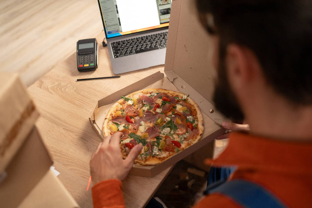 Un uomo con la pizza al computer portatile al magazzino. Ricerche sui contenuti e un tizio alla scrivania, che lavora alla startup, mangia fast food. Copia spazio - Foto, immagini