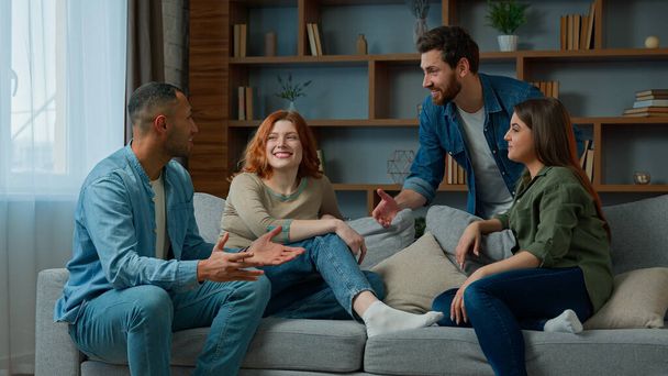 Four ethnic friends fellows companions sit on home couch talking discuss plans chatting friendly talk indoors gathered multiethnic men and women converse joking laughing sharing news real friendship - Photo, Image