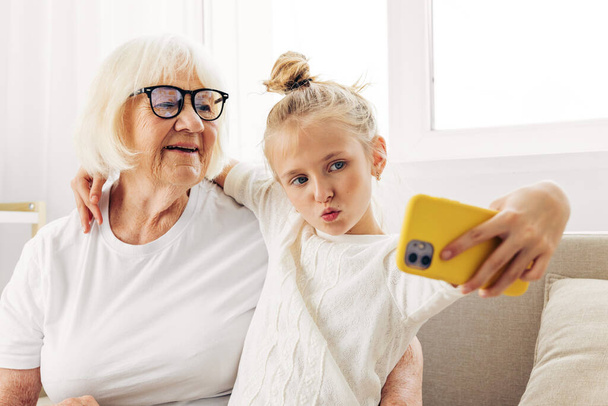 Child sofa indoors phone video copy space togetherness bonding grandmother family granddaughter photography call selfie t-shirt white education smiling hugging - Photo, Image