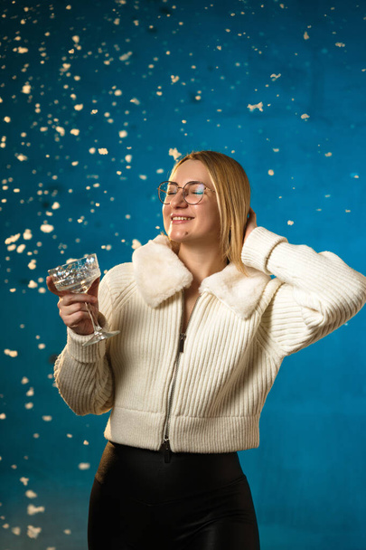Mujer rubia linda caucásica en suéter de piel blanca sobre fondo azul. Nieve Falsa fluye sobre ella. Ella es feliz, celebrando la Navidad y el año nuevo. Retrato de estudio - Foto, imagen