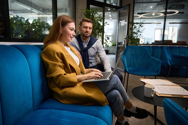 Woman and man are working with documents in coworking space on a soft blue sofa, the woman has a laptop - Фото, изображение