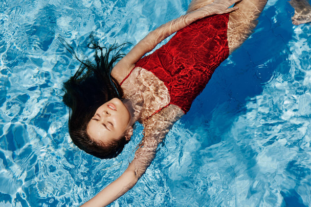 Młoda kobieta w basenie w czerwonym stroju kąpielowym z pięknym uśmiechem leżącym na wodzie i pływającym w słońcu pływającym w basenie, koncepcja relaksu na wakacjach. Wysokiej jakości zdjęcie - Zdjęcie, obraz