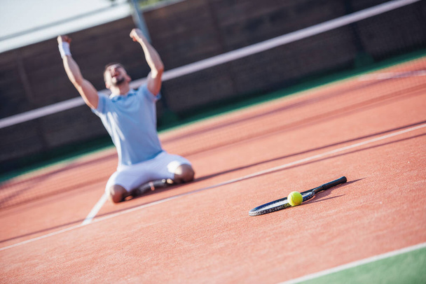 Hombre guapo está de rodillas y gritando de felicidad mientras juega al tenis en la cancha al aire libre
 - Foto, Imagen
