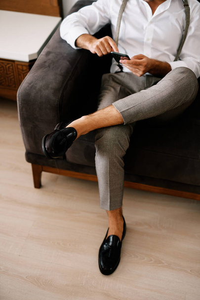 Groom clica em um smartphone em suas mãos enquanto está sentado no sofá. Cortada. Sem rosto. Foto de alta qualidade - Foto, Imagem
