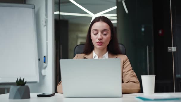 Egy álomszép kaukázusi üzletasszony portréja, aki a modern laptop előtt ül és gondolkodik. Női irodai alkalmazott elegáns öltönyben, aki könnyed mosollyal néz félre. A technológia fogalma. - Felvétel, videó