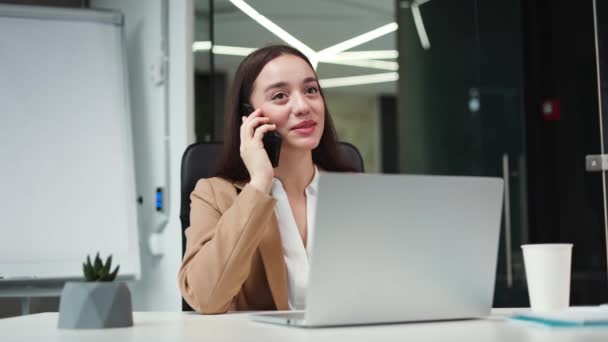 Successful female manager responding to interlocutor while talking on modern smartphone in personal office cabinet. Attractive caucasian businesswoman having conversation during workflow indoors. - Footage, Video