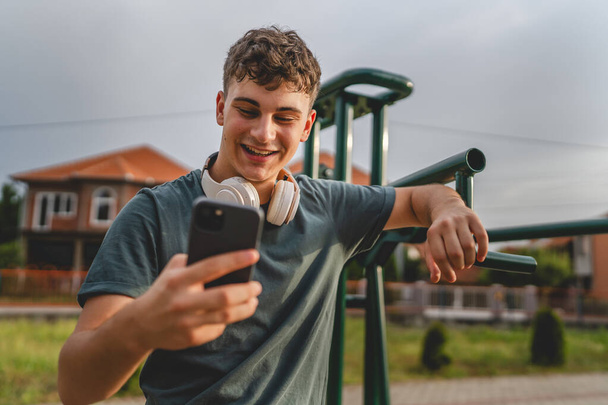 Yksi mies valkoihoinen nuori mies seistä ulkona avoin harjoituspuisto kuntosali käyttää matkapuhelin älypuhelin kuulokkeet lähettää viestejä tekstiviesti tai selata internet online-sovellus koulutukseen todellinen henkilö kopioi tilaa - Valokuva, kuva