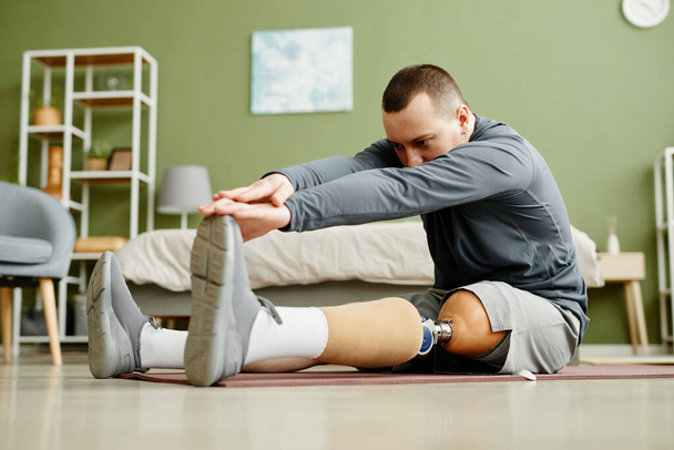 Ganzkörperporträt eines Mannes mit Beinprothese, der zu Hause Dehnübungen macht und die Zehen erreicht - Foto, Bild