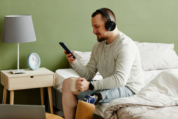 Side view portrait of smiling man with prosthetic leg enjoying morning at home and listening to music with headphones, copy space - Photo, image