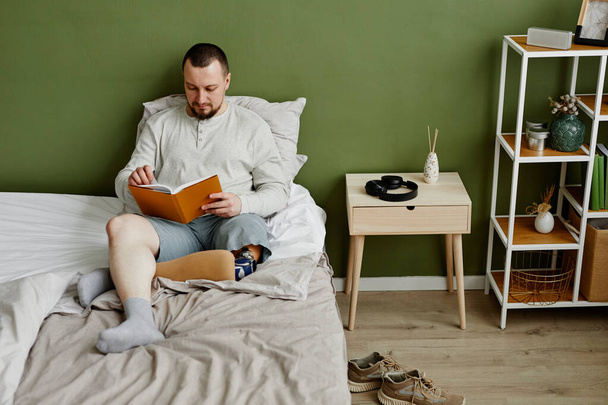 Blick aus der Vogelperspektive auf einen jungen Mann mit Beinprothese, der im Bett ein Buch liest, während er es sich zu Hause gemütlich macht, Kopierraum - Foto, Bild