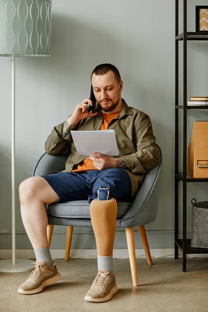 Retrato completo del hombre con la pierna protésica llamando por teléfono mientras está sentado en la silla en el interior del hogar - Foto, imagen