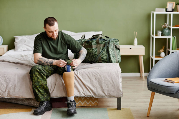 Retrato completo de veterano militar con pierna protésica que se pone uniforme del ejército en el dormitorio, espacio de copia - Foto, Imagen