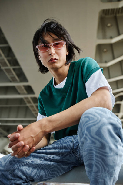 Vertikales Porträt eines asiatischen Mannes im Straßenstil, der im Freien in die Kamera schaut und eine rosa Sonnenbrille trägt - Foto, Bild