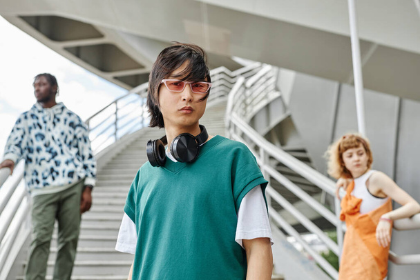 屋外でストリートスタイルの服を着ている若者の多様なグループは,前景にヘッドフォンを持つアジア人男性に焦点を当てます - 写真・画像