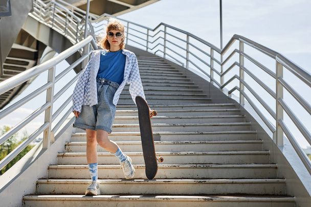 Retrato de comprimento total da jovem com skate posando ao ar livre sob a luz do sol e em pé sobre escadas, espaço cópia - Foto, Imagem