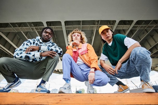 Grupa trzech młodych ludzi w ulicznym stylu ubrania na zewnątrz podczas siedzenia na schodach w obszarze miejskim i patrząc w kamerze - Zdjęcie, obraz