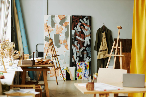 Parte de un amplio estudio de artes con caballetes de madera, pinturas sobre lienzos, ropa de trabajo colgando en la pared y otros suministros y equipos - Foto, Imagen