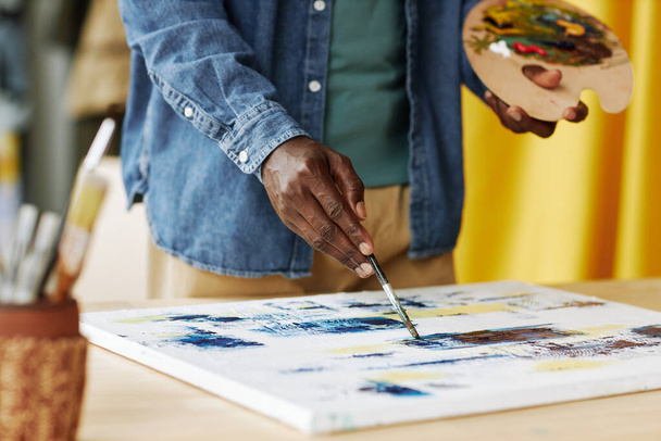 Hand van jonge Afro-Amerikaanse mannelijke kunstenaar met kleurenpalet schilderen met acrylverf op doek terwijl de stand-by werkplek - Foto, afbeelding