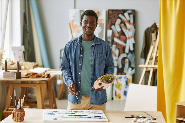 Молодой успешный афроамериканец в повседневной одежде держит деревянную цветовую палитру с акриловыми красками при создании произведений искусства - Фото, изображение