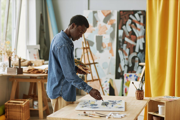 Joven hombre negro creativo con pincel y paleta de colores trabajando sobre nuevas obras de arte pintadas con pinturas acrílicas por lugar de trabajo en el estudio - Foto, Imagen