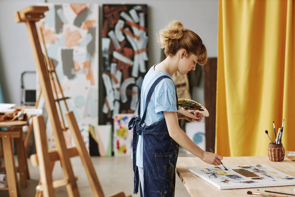 Вид сбоку на молодую блондинку в джинсовом фартуке, рисующую картины акриловыми красками на рабочем месте с холстом - Фото, изображение