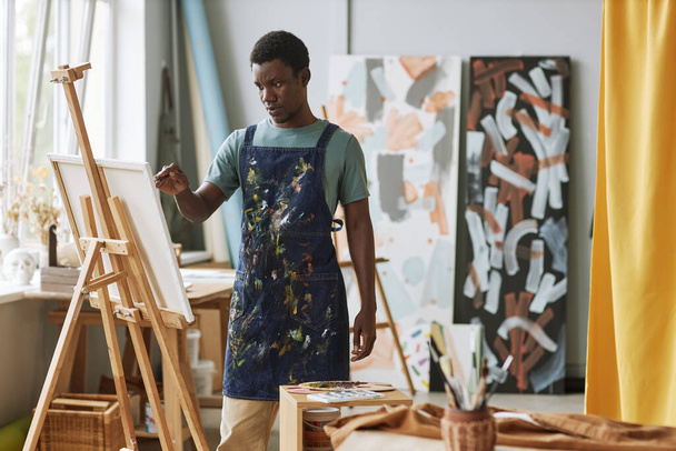 Νεαρός σοβαρός Αφρο-Αμερικανός άνδρας με ποδιά denim στέκεται μπροστά από καμβά στο καβαλέτο και δημιουργεί νέα ζωγραφική στο lesiure - Φωτογραφία, εικόνα