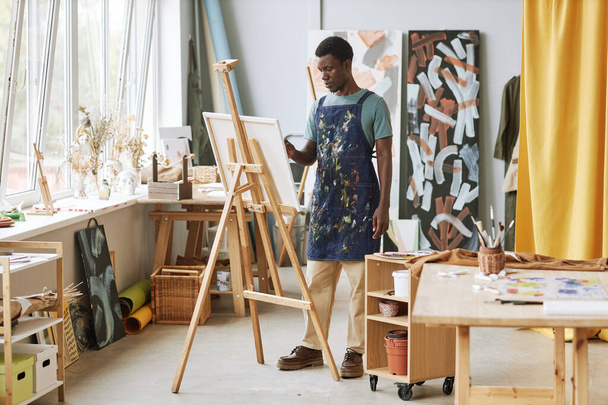 Молодой серьезный творческий мужчина в фартуке стоит перед мольбертом с незавершенными произведениями искусства во время работы над новой картиной в студии - Фото, изображение