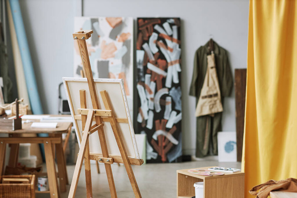 Worplace of modern artist with canvas on wooden easel standing in the center of spacious studio or classroom of school of arts - Photo, Image