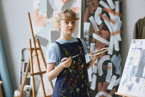 Mladý pilný student umělecké školy s barevnou paletou a štětcem stojící před kamerou a stojanem s novým mistrovským dílem - Fotografie, Obrázek