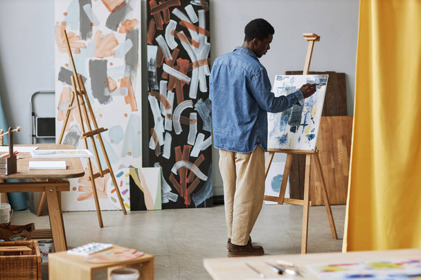 Πίσω από το νεαρό μαύρο άνδρα σε casualwear εργάζονται πάνω σε νέα ζωγραφική, ενώ στέκεται μπροστά από easel με καμβά στο μάθημα στο σχολείο των τεχνών - Φωτογραφία, εικόνα