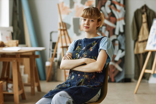 Νεαρή ξανθιά καλλιτέχνιδα ή δασκάλα ζωγραφικής που κάθεται σε καρέκλα στο στούντιο ή στην τάξη της σχολής τεχνών με σταυρωμένα τα χέρια στο στήθος - Φωτογραφία, εικόνα