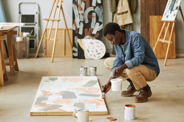 Young black man sitting on squats and bending over canvas with unfinished artwork while painting it in studio or classroom - Photo, Image