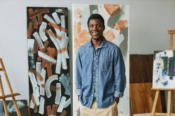 スタジオや教室での抽象絵画の背景に立っているカジュアルウェアでアフリカ系アメリカ人の若い笑顔 - 写真・画像