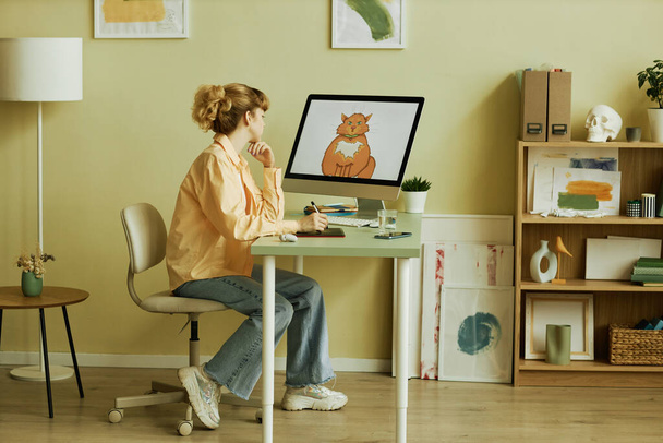 Joven artista gráfica rubia creativa en ropa casual sentada en el lugar de trabajo frente a la pantalla de la computadora con imagen dibujada de gato - Foto, imagen