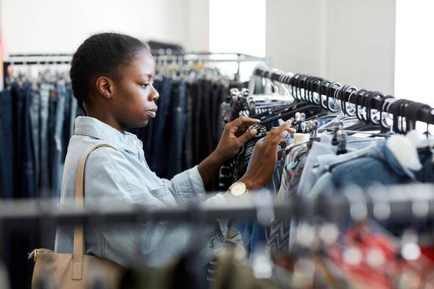 Retrato de vista lateral de una joven mujer negra mirando la ropa en un estante en una tienda de segunda mano - Foto, Imagen