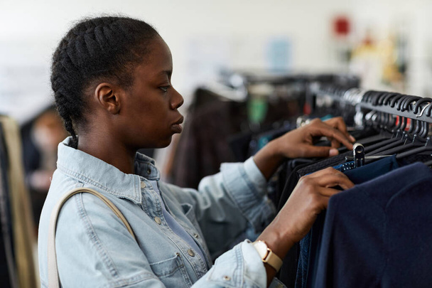漂流店で買い物しながらラックで服をブラウジングする黒人の若い女性の側面の肖像画 - 写真・画像