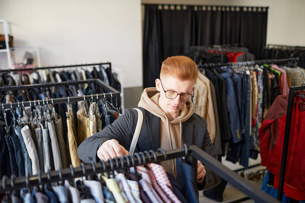 Portrait grand angle de jeune homme roux regardant des vêtements tout en faisant des achats durables dans un magasin d'épargne - Photo, image
