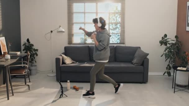 Full shot of woman wearing headphones and sportswear air boxing with dumbbells while standing in her living room - Footage, Video